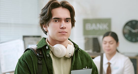 Portrait of Caucasian young adult man standing in bank branch, holding clipboard and wearing headphones around neck with bank employee working at counter in background